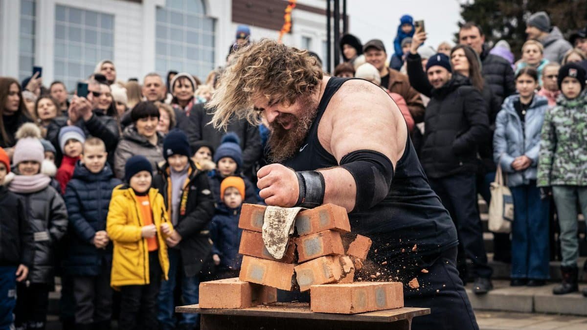 Strongman breaks bricks in front of a cheering crowd outdoors, showcasing strength and performance.