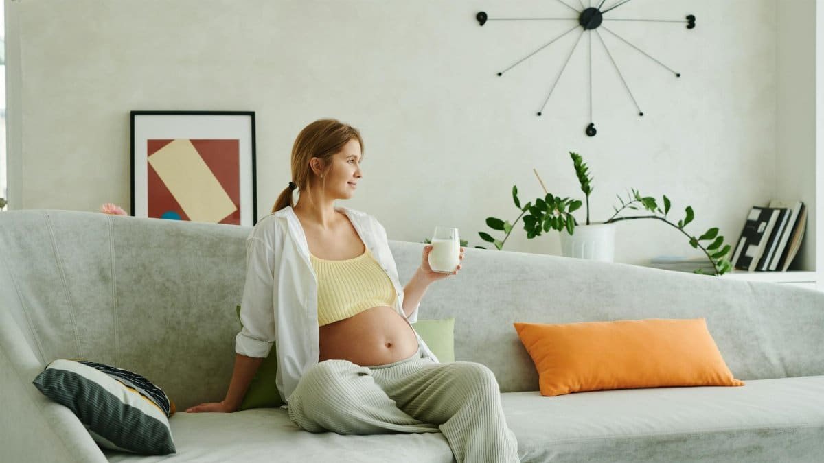 Expectant woman sitting on a sofa indoors, holding a glass of milk, smiling, and enjoying a peaceful moment.