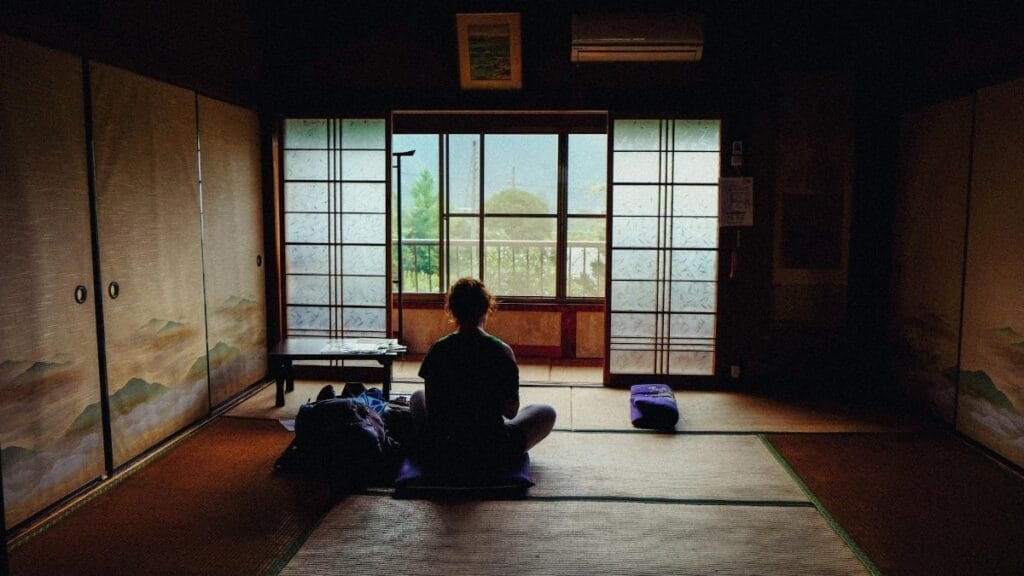 A person meditating in a tranquil traditional Japanese room, facing the view outside.