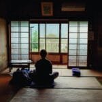A person meditating in a tranquil traditional Japanese room, facing the view outside.