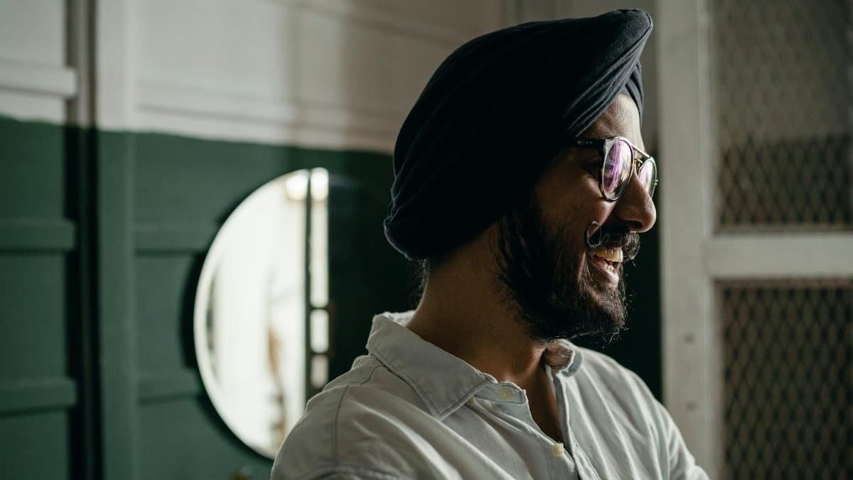 Side view of bearded positive laughing Indian male in eyeglasses wearing casual shirt and turban standing near green and white wall and mirror and looking away happily
