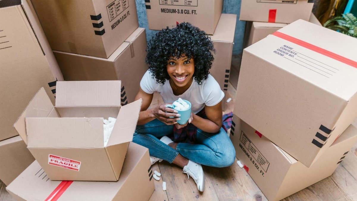 A woman sitting among moving boxes with a mug, surrounded by packing materials.