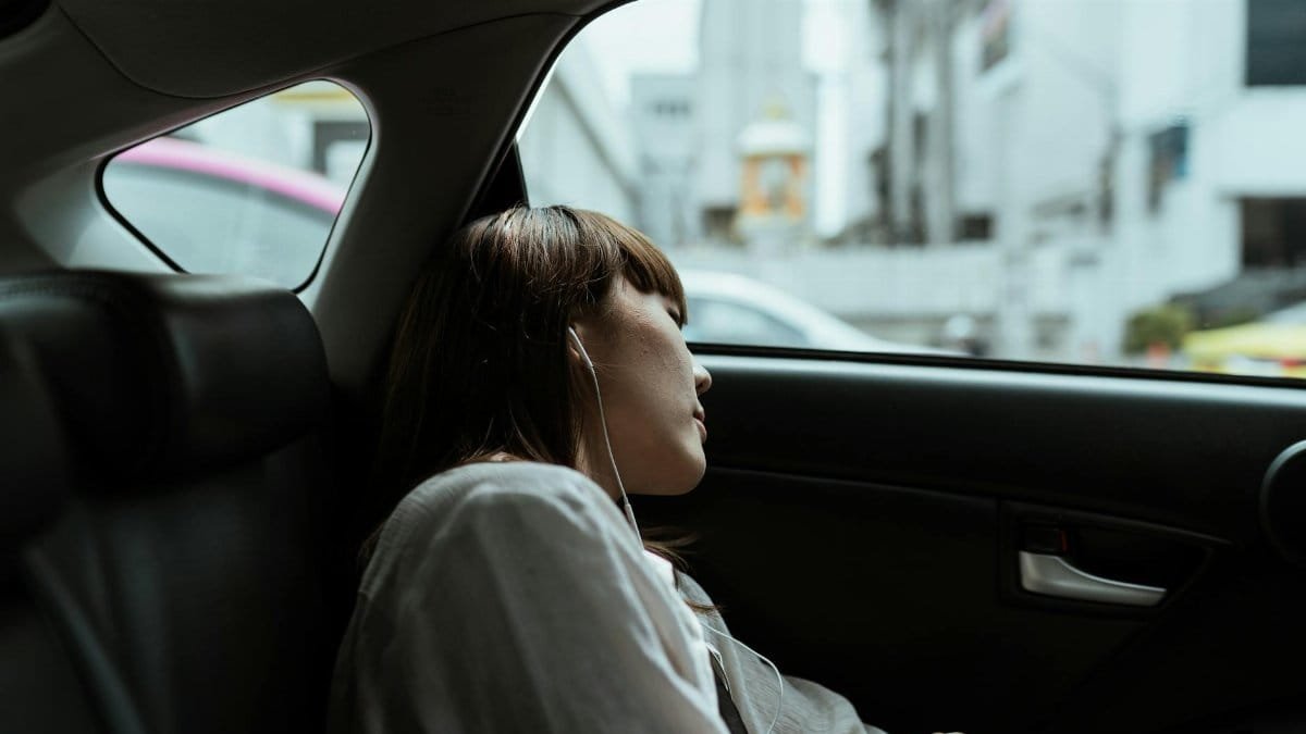 Side view of unemotional female in white shirt resting in car passenger seat and listening to music via earphones while riding in urban environment