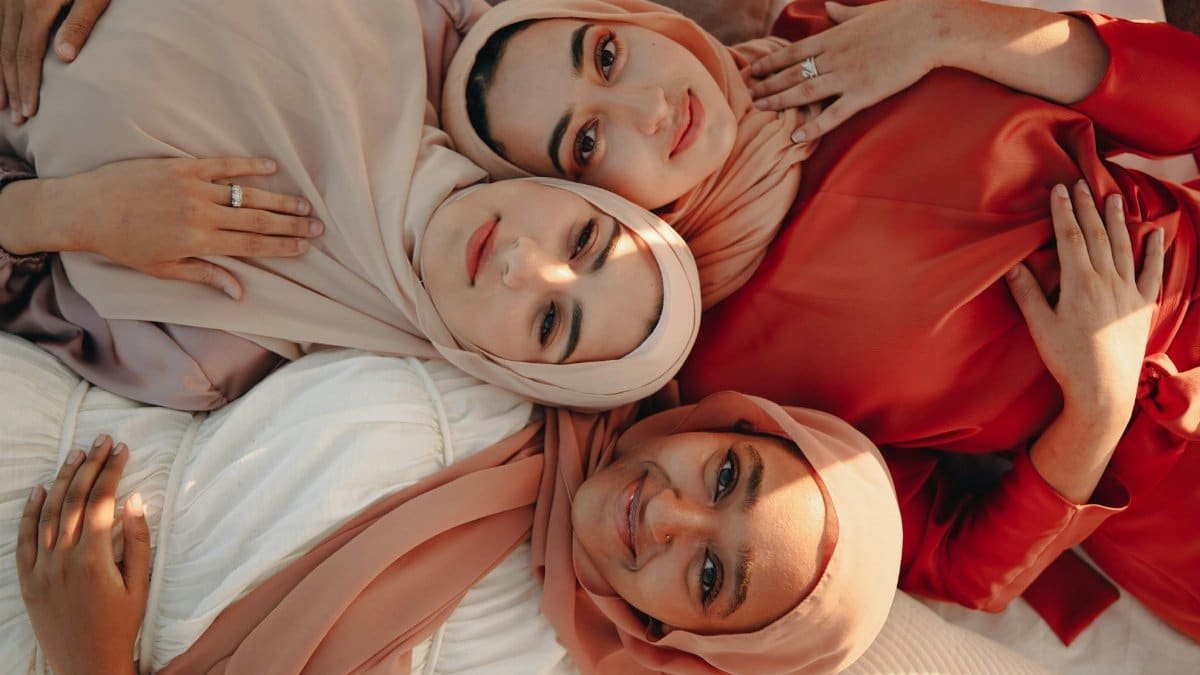 Three women in hijabs, dressed in warm tones, lying together and embracing.