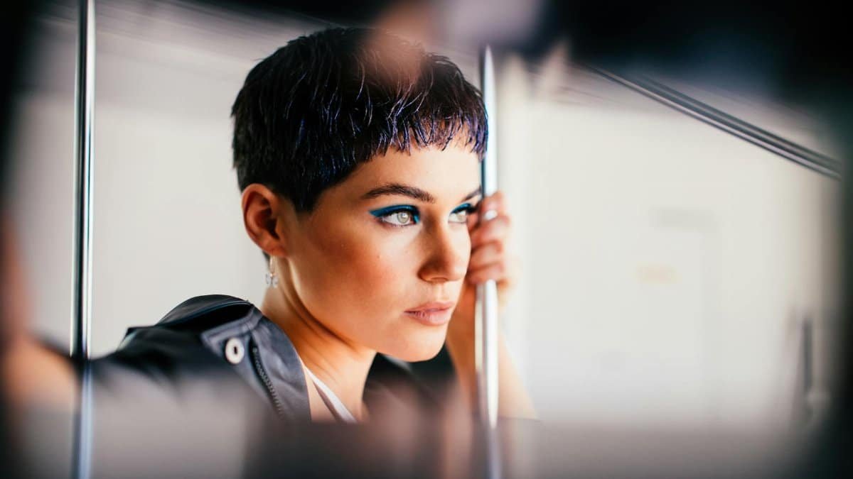 A young woman with short dark hair and bold makeup ponders while holding a railing in public transport.
