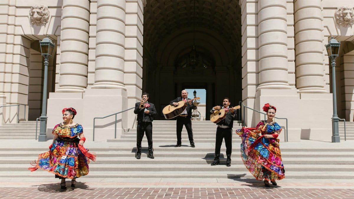 Mariachi band and dancers in traditional clothing performing outside a grand building with steps.