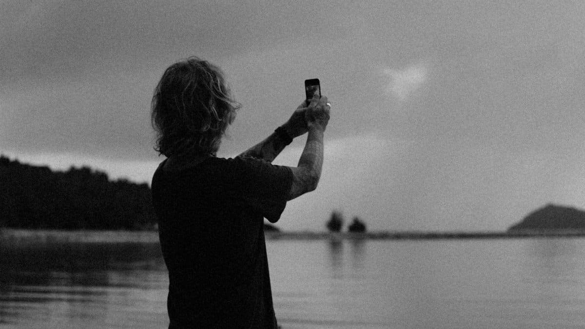 A person uses a smartphone to capture a lakeside view in a dramatic monochrome setting.