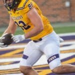 Athlete in yellow jersey scores a touchdown during a college football game outdoors.