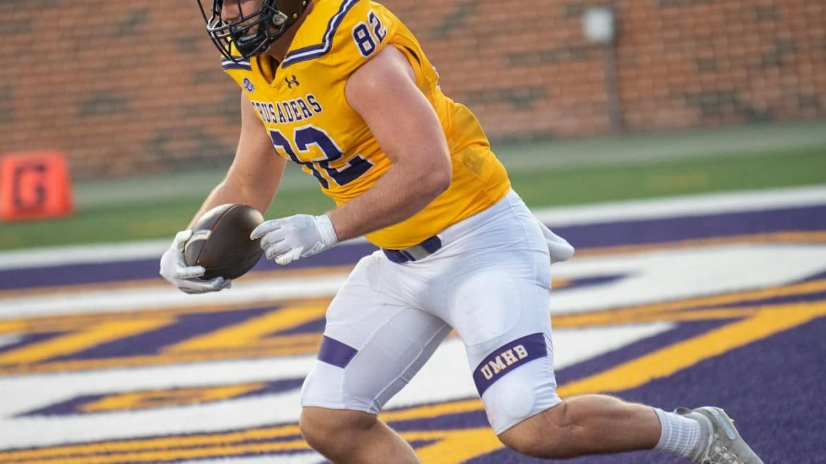 Athlete in yellow jersey scores a touchdown during a college football game outdoors.