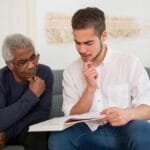 Young man and senior discussing a book at home, fostering intergenerational connection.