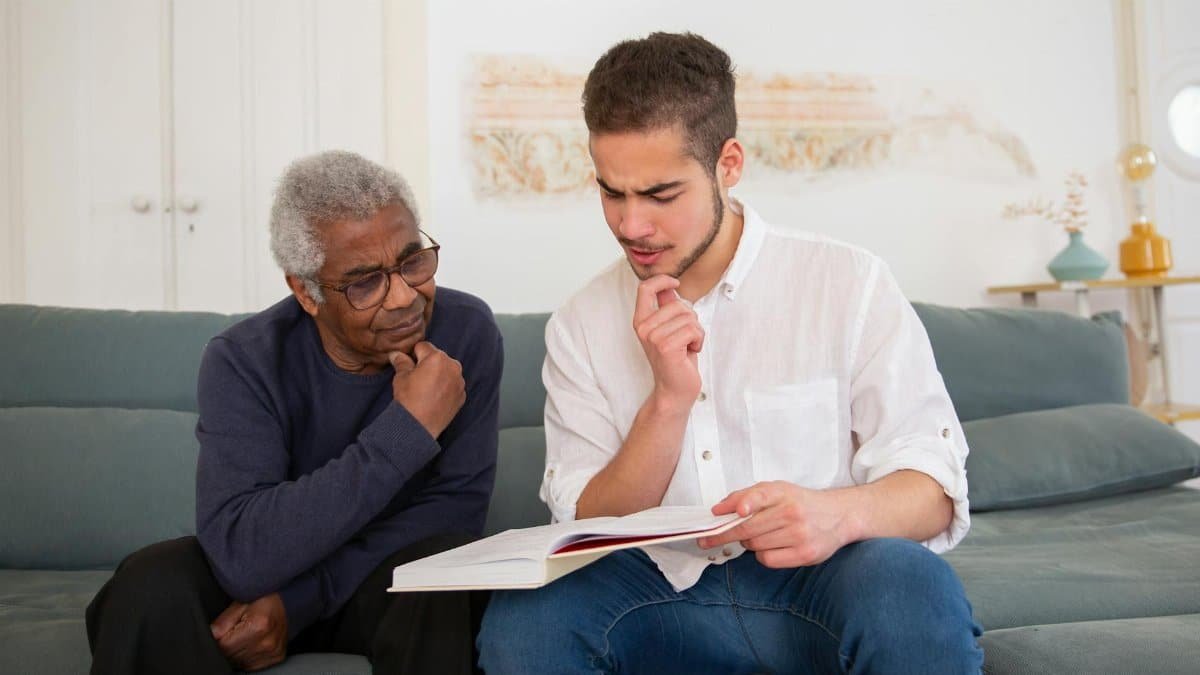 Young man and senior discussing a book at home, fostering intergenerational connection.