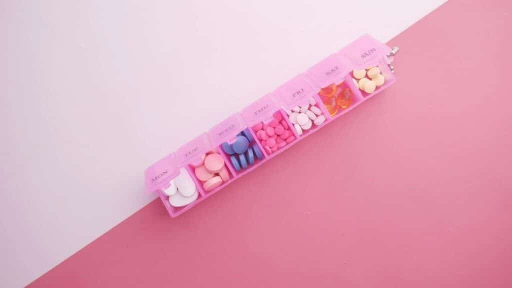 A pink pill organizer with colorful tablets arranged for each day of the week on a vibrant pink background.