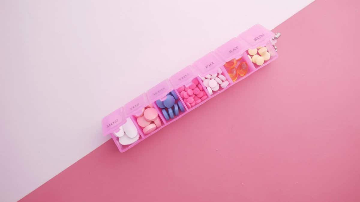 A pink pill organizer with colorful tablets arranged for each day of the week on a vibrant pink background.