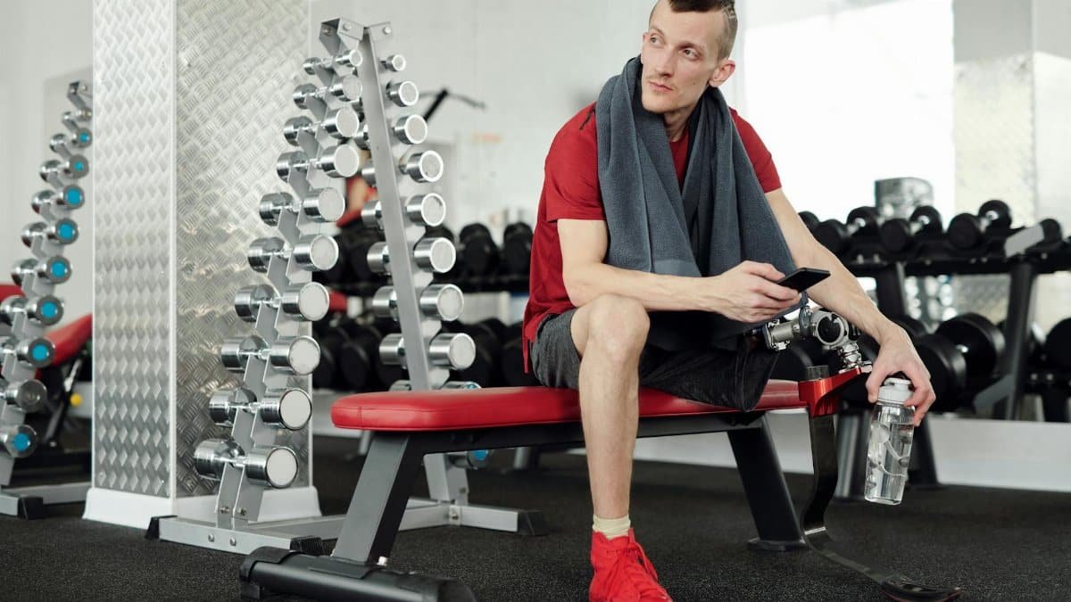 Man with prosthetic leg taking a break in gym, holding smartphone and water bottle.