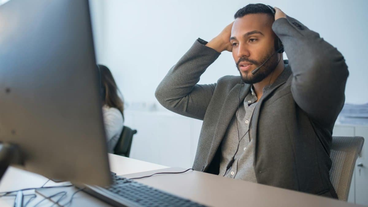Man in a headset working at a desk, hands on head, conveys work stress.