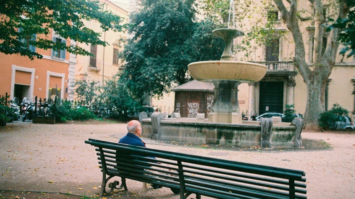 Elderly man sitting on a park bench near a fountain, enjoying a quiet urban setting.