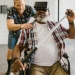 Man assists wheelchair user engaged in rehabilitation exercise with VR set indoors.