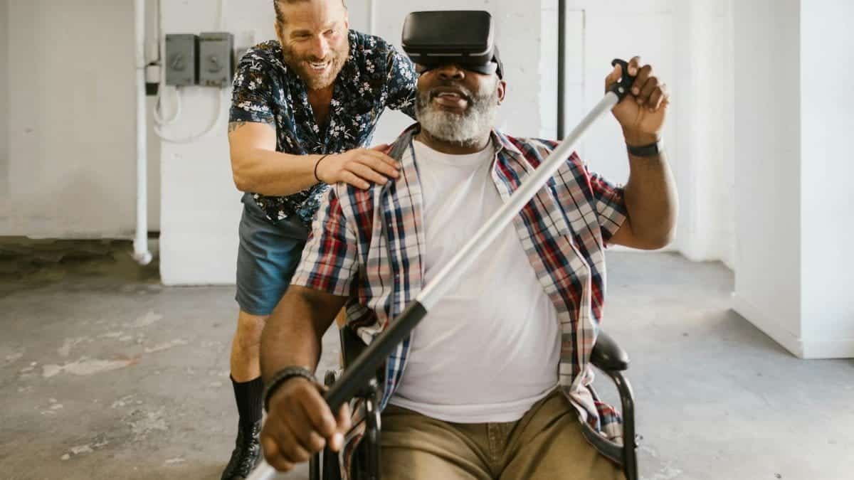 Man assists wheelchair user engaged in rehabilitation exercise with VR set indoors.