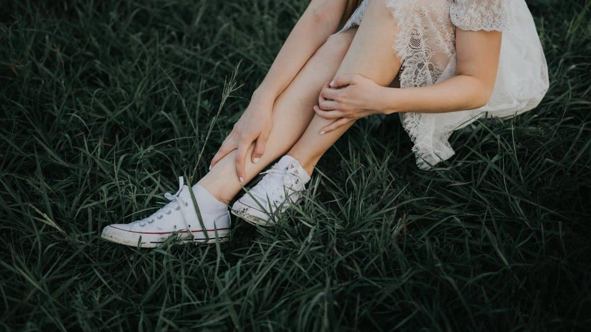 A woman in a white lace dress sitting on green grass wearing white sneakers.