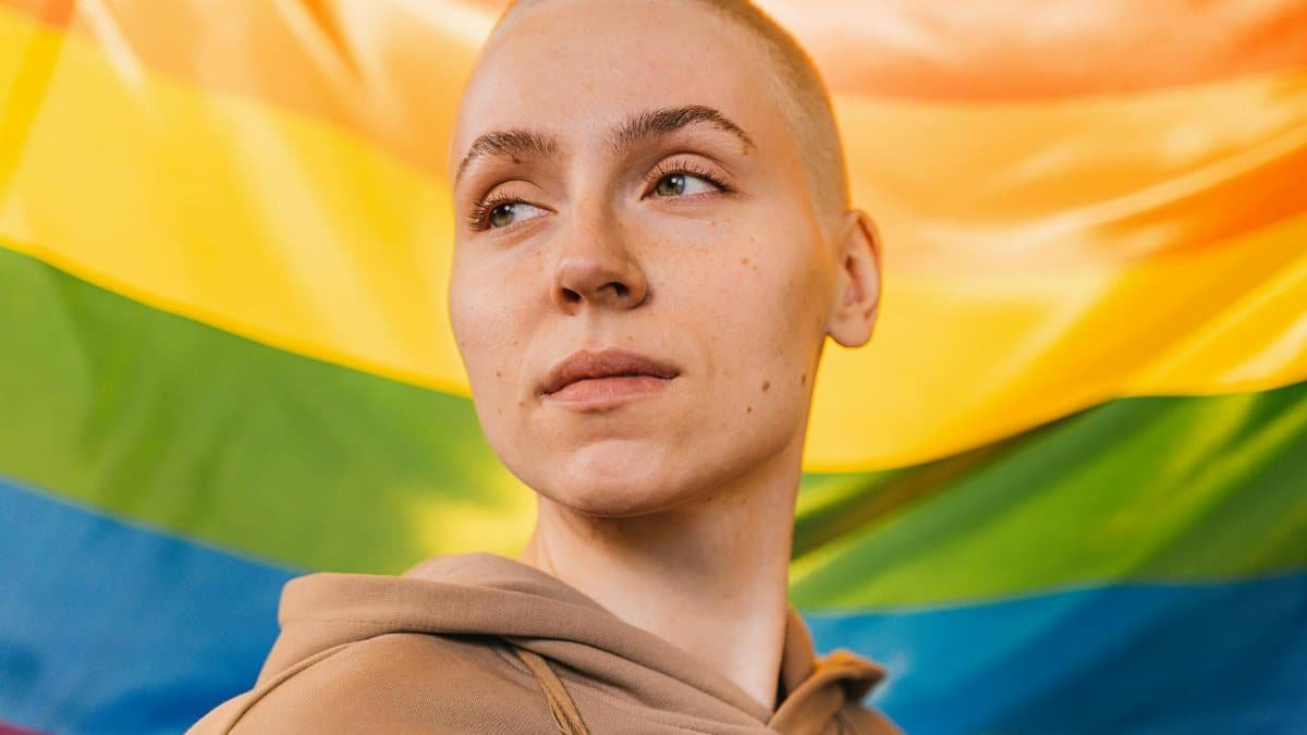 Close-up portrait of an adult with a rainbow flag backdrop, symbolizing pride and diversity.