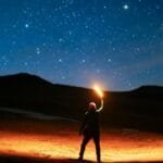 A lone hiker holding a lantern illuminates the night sky filled with stars, creating a magical scene.