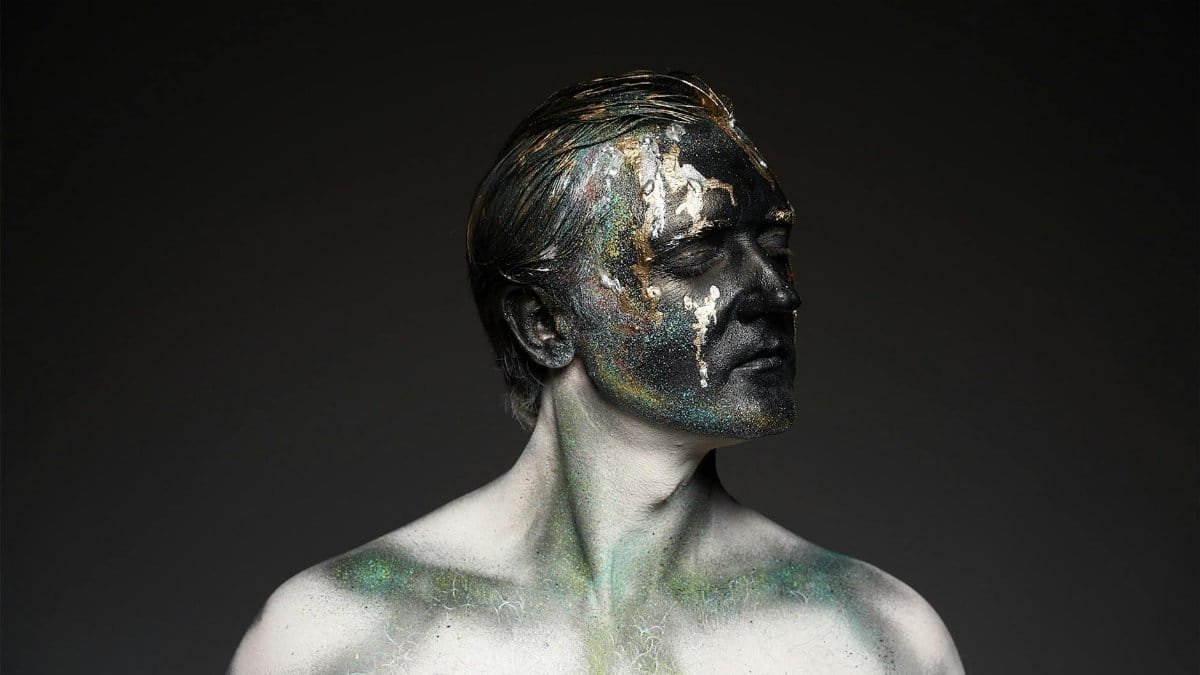 Male shirtless model with glowing glitters on black painted face covering eyes standing on black backdrop