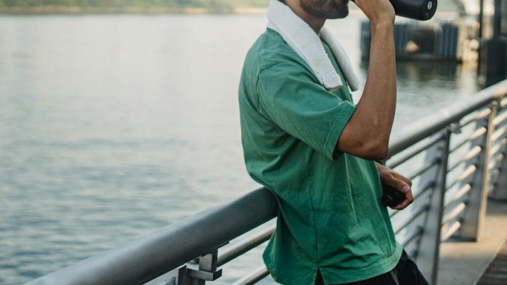 A man in activewear drinks water by the river, embracing a healthy lifestyle.