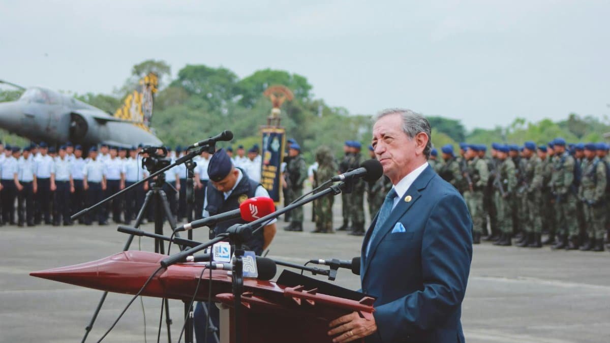 Side view of mature colonel with order standing near microphones on asphalt pavement behind army of soldiers near airplane monument under serene sky during national military holiday and looking away