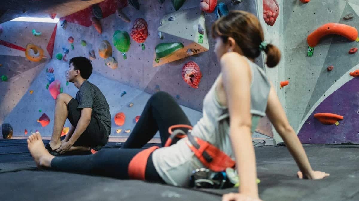 Two young adults resting on mats in an indoor bouldering gym, surrounded by colorful climbing holds.