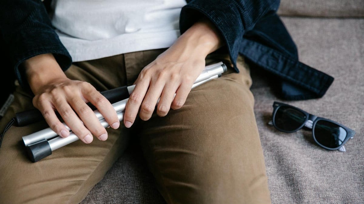Close-up of a person holding a white cane with sunglasses on a sofa. Emphasizes accessibility.