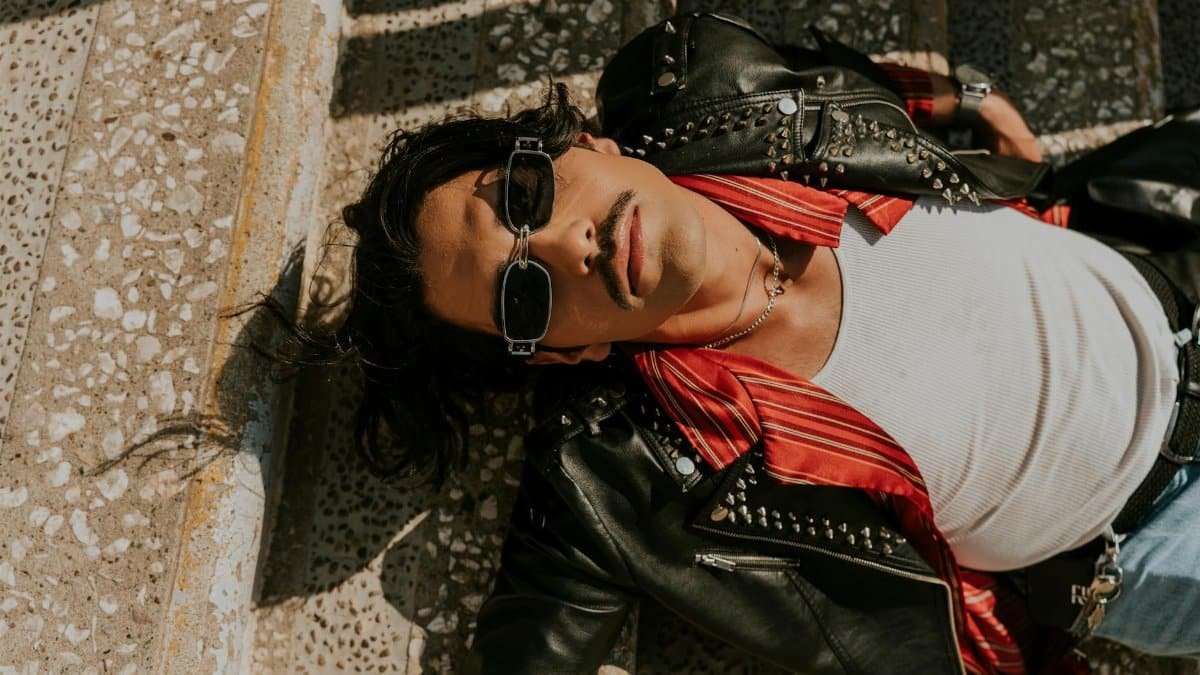 A fashionable man in a leather jacket relaxes on stone steps under the sun.