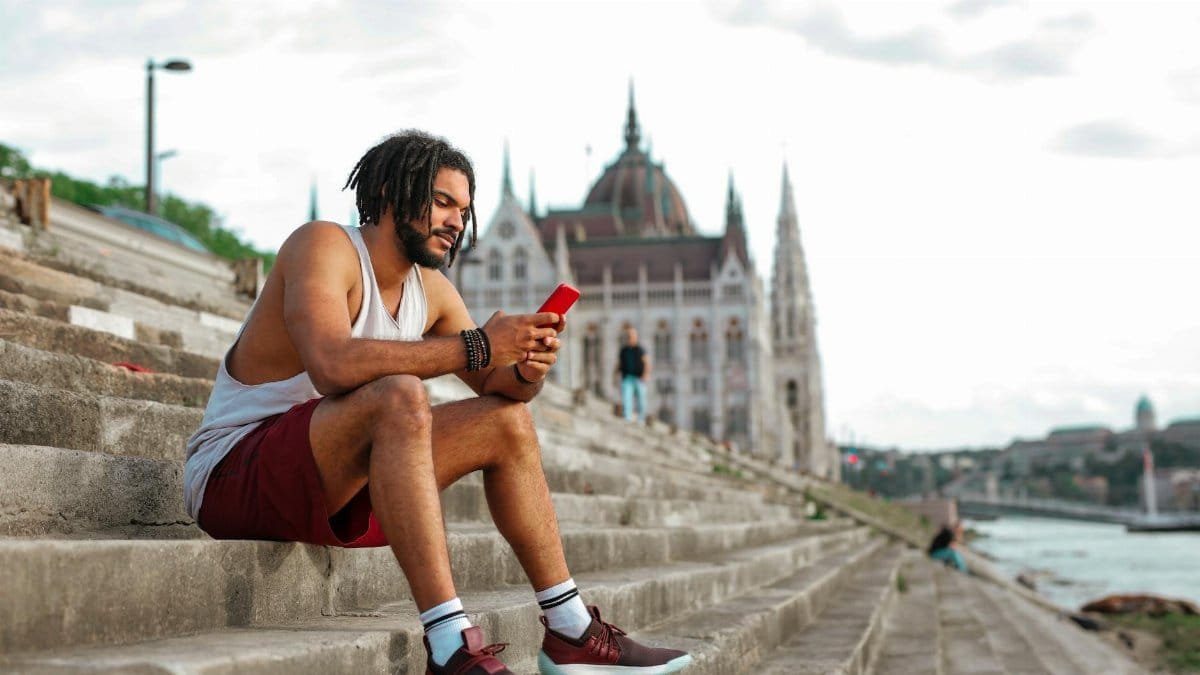 Young man sits casually on riverside steps in Budapest, engaged with his phone. Urban summer scene.