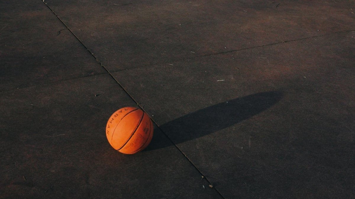 A single basketball casting a shadow on a textured outdoor court surface at dusk.