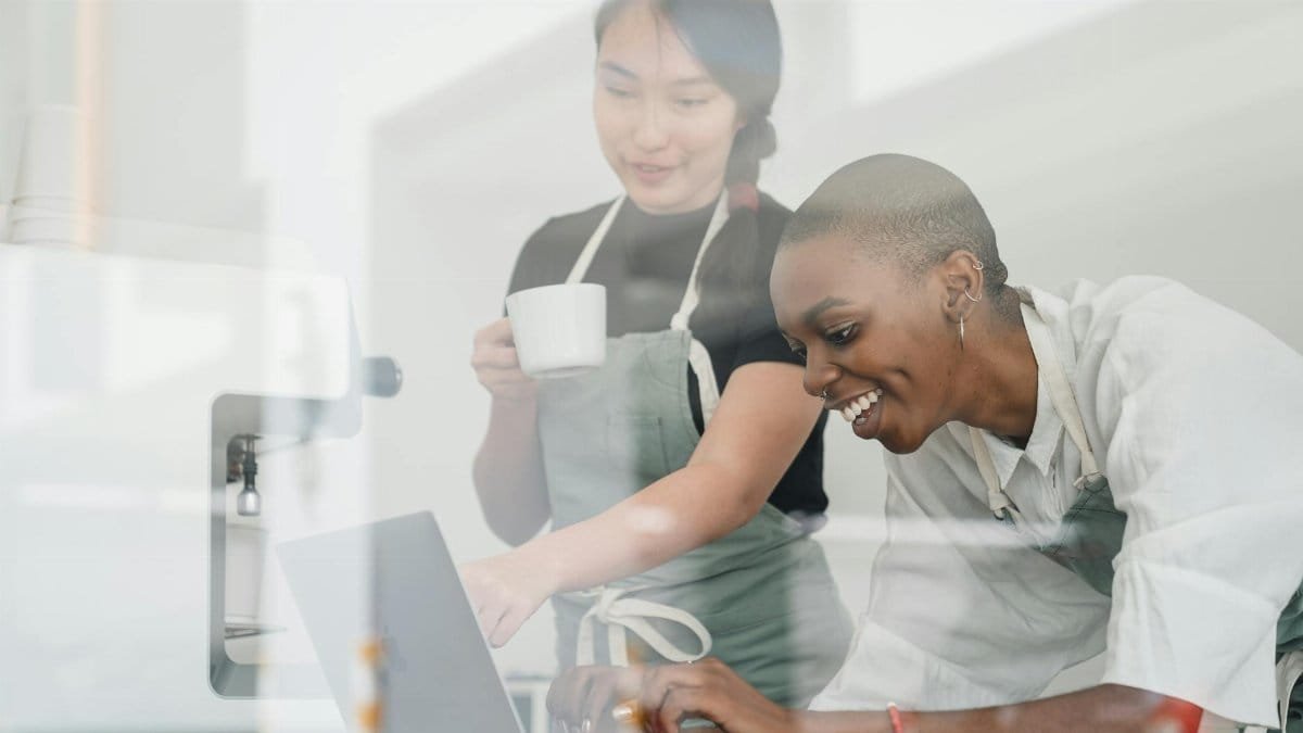 Through glass of friendly young Asian head barista showing young cheerful African American female coworker how to use special program on laptop