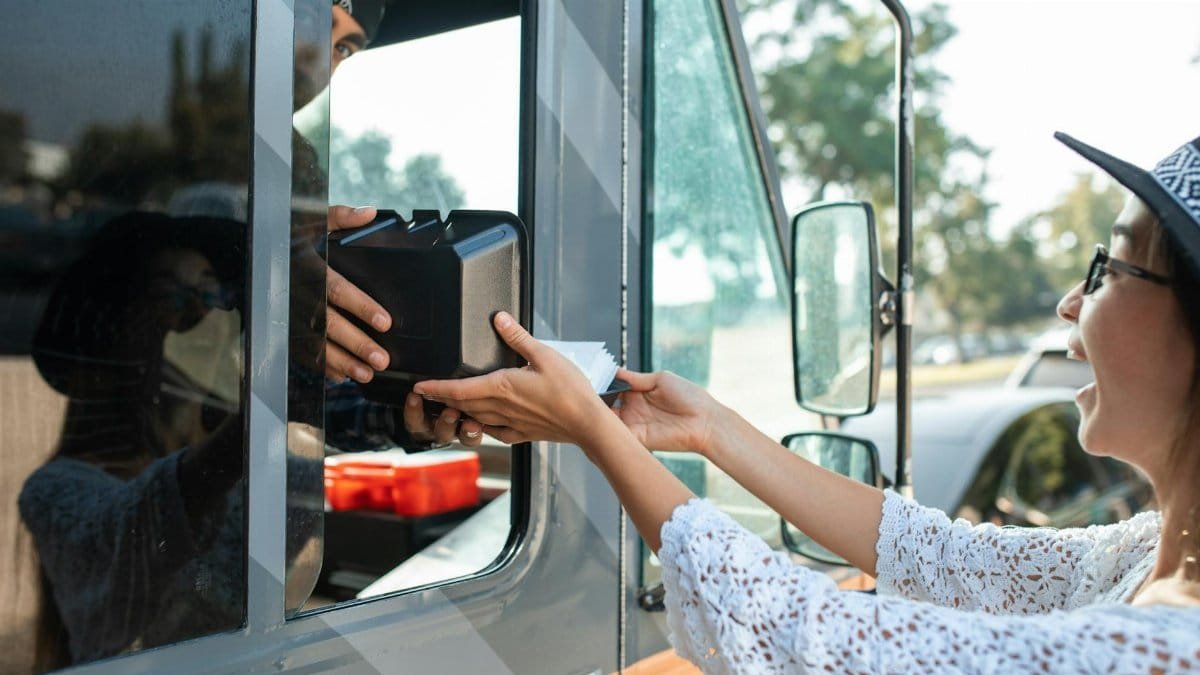A joyful woman receives a boxed meal from a food truck window on a sunny day.