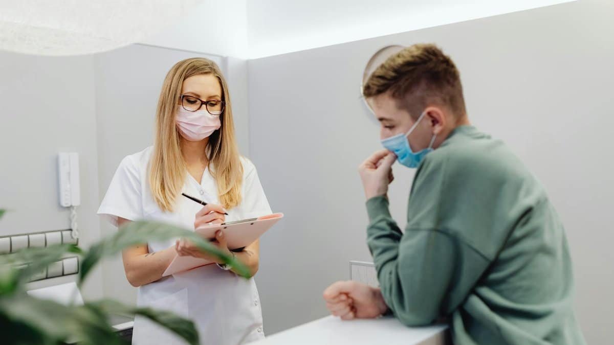 A healthcare worker consults a patient at a clinic reception, both wearing face masks.