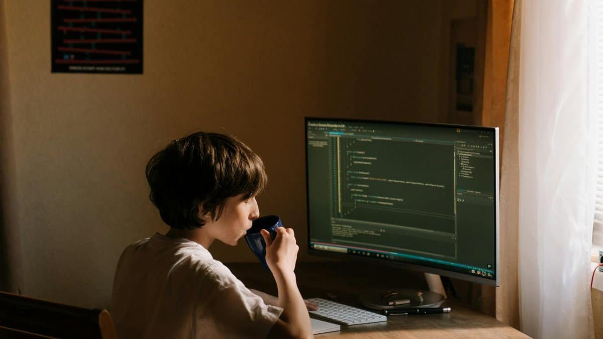 A young person coding at a desk with a computer and drinking from a mug.