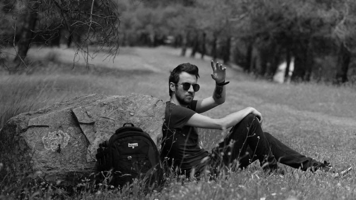 A man relaxes on a grassy hillside with a backpack and raised hand, in a serene outdoor setting.