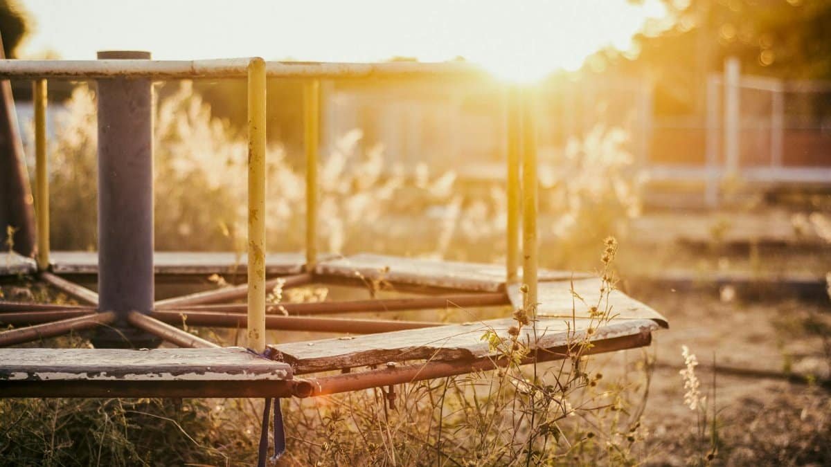 A sunlit, rusted merry-go-round in an abandoned playground overgrown with grass.