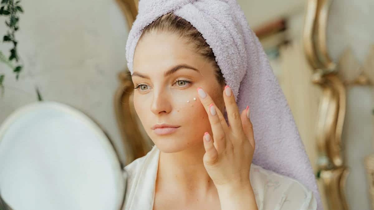 Woman with head towel applying moisturizer in front of mirror indoors.