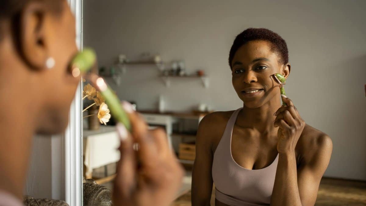 A woman enjoying her skincare routine using a jade roller in front of a mirror.