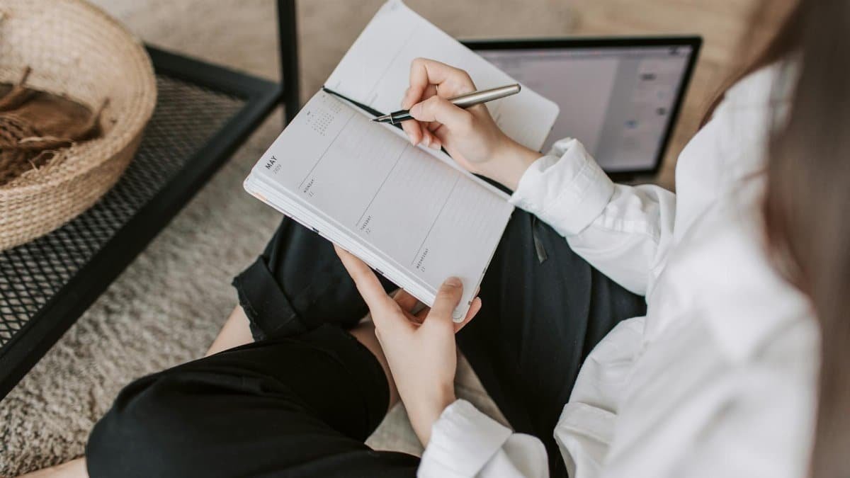 Side view of faceless woman in casual clothes taking notes on notepad while sitting in lotus pose on floor in modern apartment during daytime