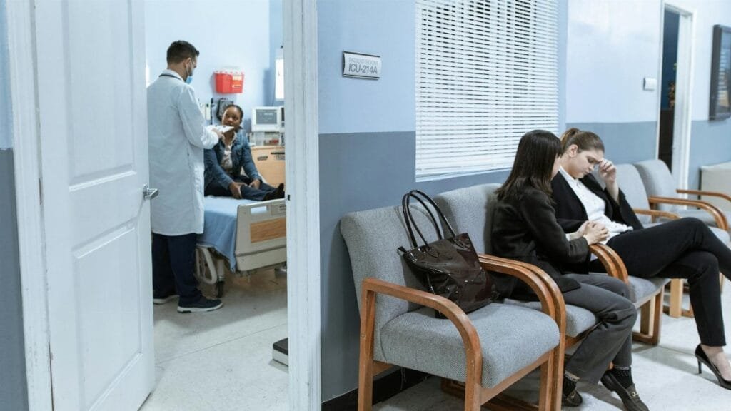 Doctor with patient in ICU, women in waiting area showing concern.