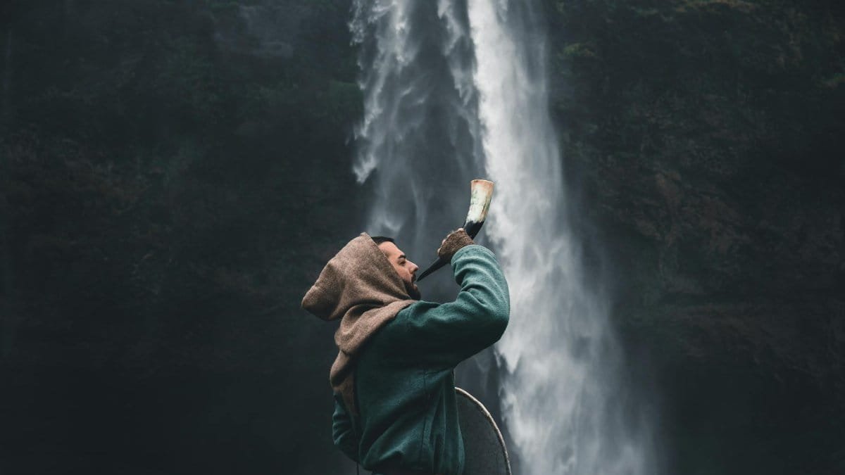 A warrior in a hooded cloak standing by a waterfall in Iceland, holding a horn.