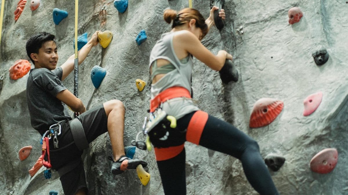 Low angle of young Asian male and female friends in sportswear and protective harness practicing bouldering together in climbing gym