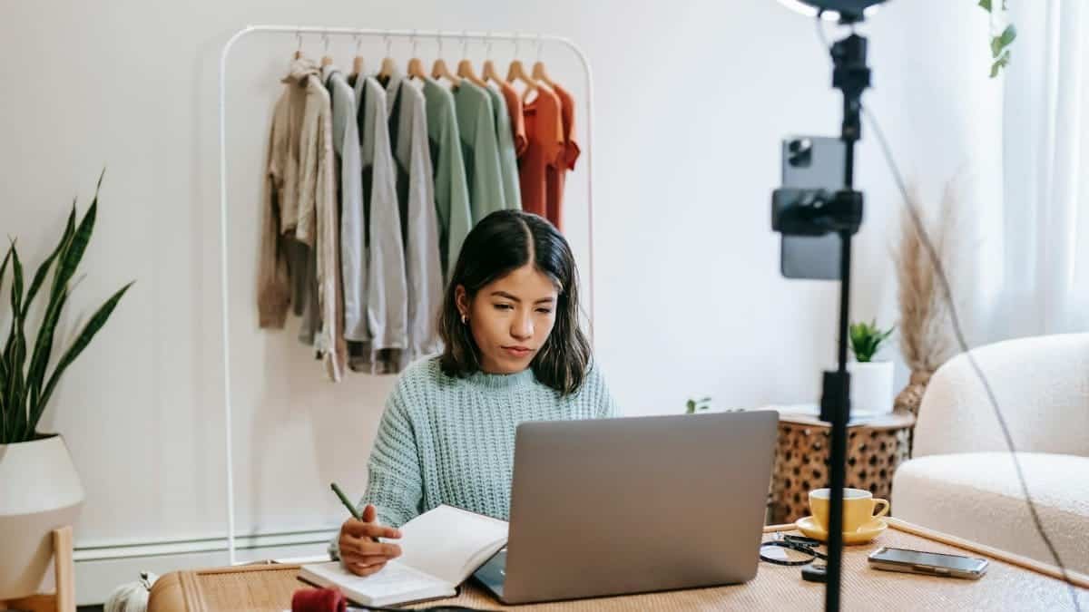Focused young woman working on laptop in home office, surrounded by clothing rack and camera setup.