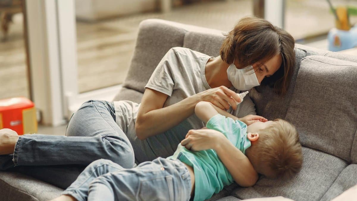 A mother wearing a face mask comforts her sick son with nasal drops on the sofa.