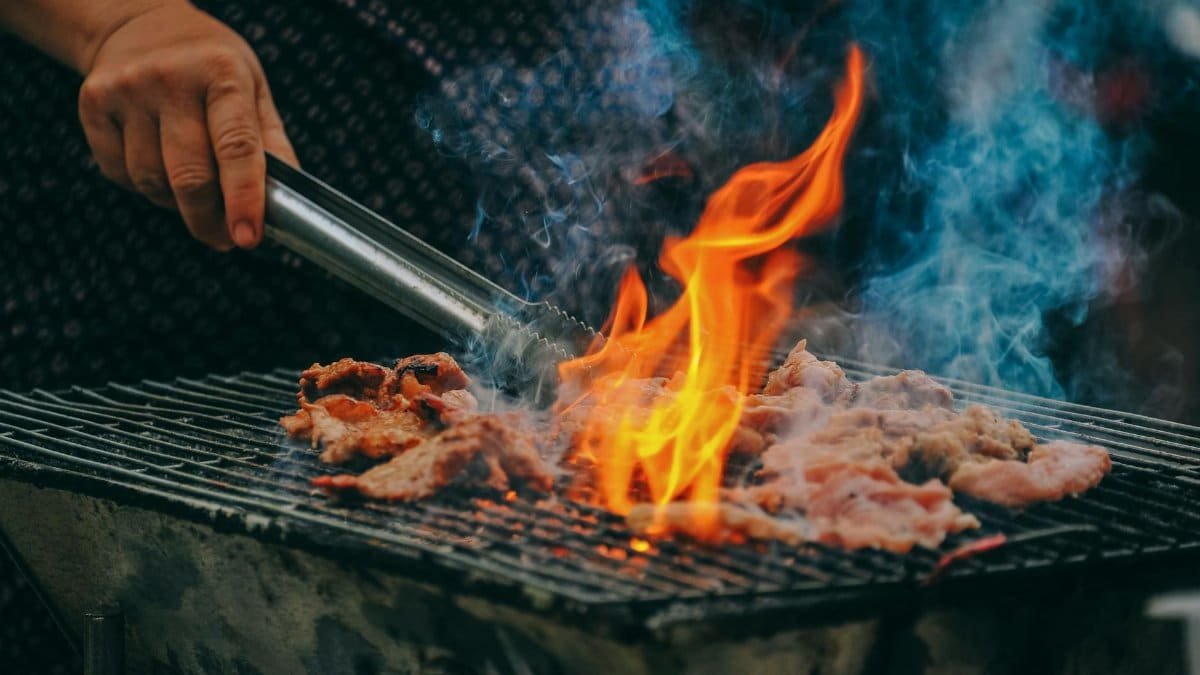Close-up of grilling meat with vibrant flames and smoky aura, showcasing outdoor cooking.