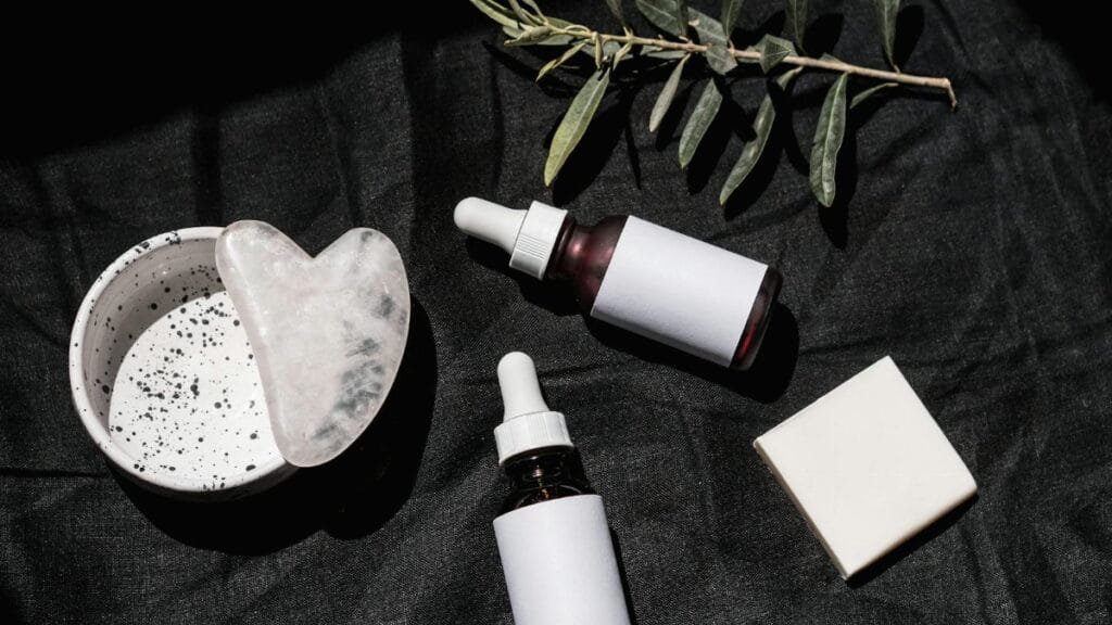 Flat lay of skincare products with gua sha and leaves on black textile. Ideal for beauty and wellness visuals.