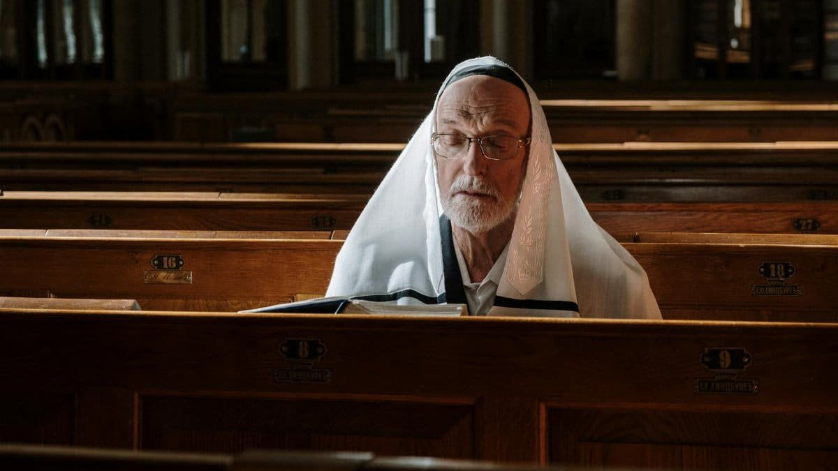 An elderly man sits in a synagogue pew while wearing a tallit and praying with a book.